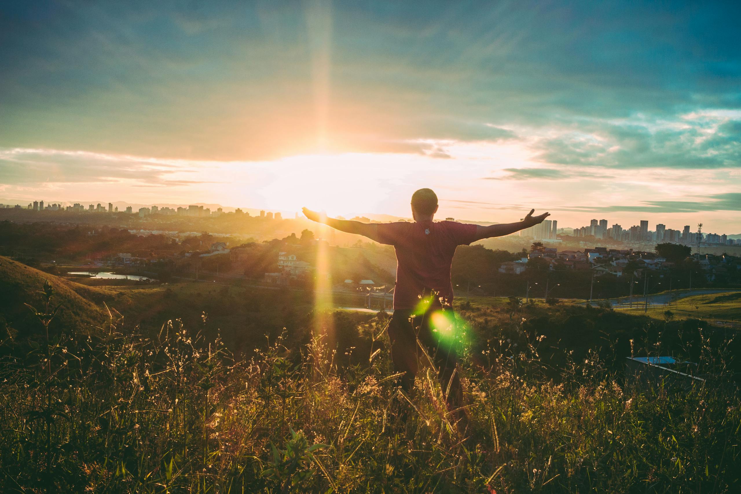 Person standing on grassy hill with open arms embracing a vibrant sunset over a cityscape.